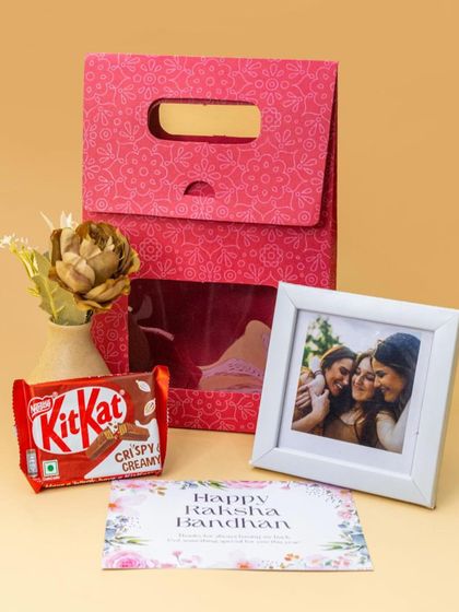 A lovely Rakhi gift set for a sister. It comes with a personalized photo frame, a KitKat, a 'Happy Raksha Bandhan' card, and a decorative flower, all in a pretty pink gift bag.