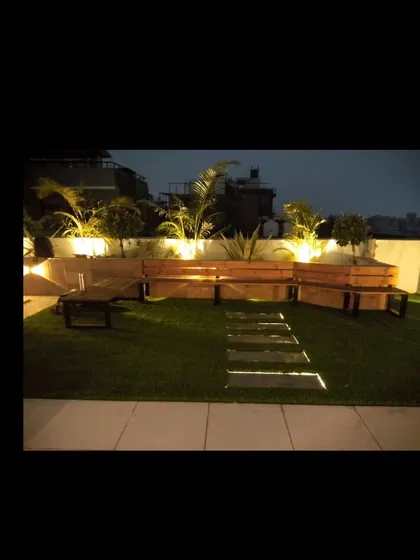 The same thermopine wood bench at night, with uplighting in the planters highlighting the palm trees and creating a relaxing ambiance.