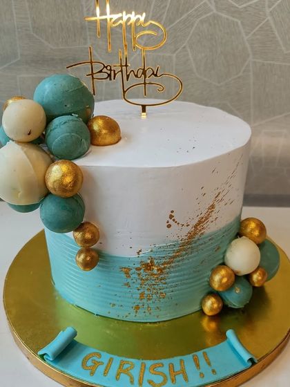 A modern and stylish cake with a teal and white color scheme. The design is accented with a splash of gold and a cascade of chocolate spheres.