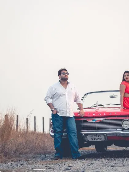 A stylishly composed shot of the couple posing with a red convertible in a rustic setting, perfect for a high-fashion pre-wedding album.
