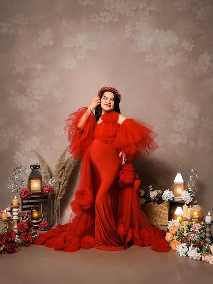 A vibrant solo portrait of the mom-to-be in a stunning red ruffled gown. She is surrounded by a lavish setup of flowers and candles, creating a rich and celebratory feel.