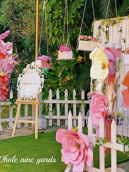 A whimsical garden party photo booth area. The giant paper flowers, white picket fence, and hanging floral baskets created a perfect spot for pictures.