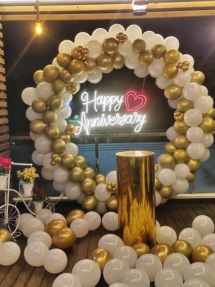 An elegant and classic anniversary decoration with a full circular arch of gold and white balloons. The "Happy Anniversary" neon sign with a heart detail adds a romantic touch, perfect for an intimate party.