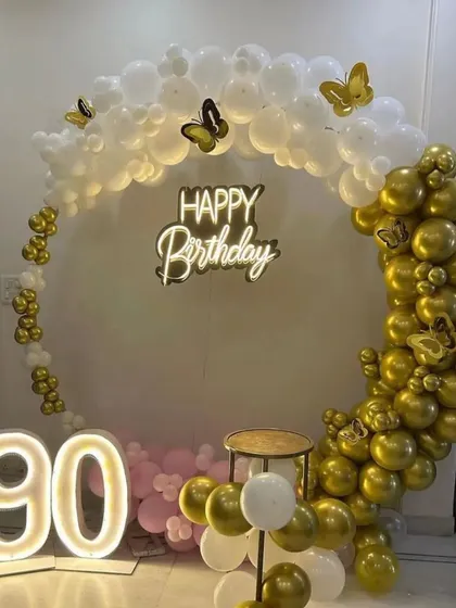 A grand 90th birthday celebration. This setup features a circular balloon arch in white, gold, and pink, a 'Happy Birthday' neon sign, and large '90' marquee numbers.