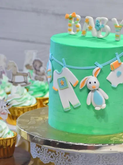 A charming baby shower cake with a clothesline theme. The cake is decorated with tiny, edible baby clothes, a bunny, and "BABY" blocks, paired with matching cupcakes for the celebration.
