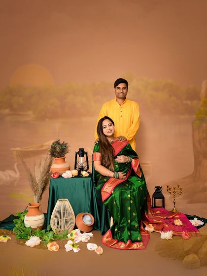 A classic portrait of an expecting couple in a traditional setting. The mom-to-be is seated in a beautiful dark green saree, creating a rich and elegant composition.