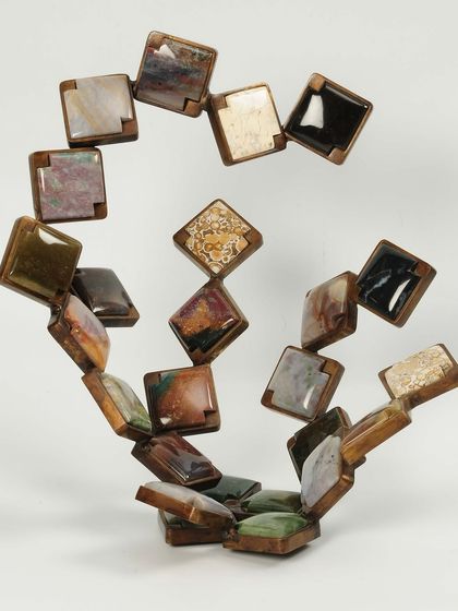 An early freestanding sculpture balancing geometric precision with organic variety, using different types of stone set in a swirling metal frame.