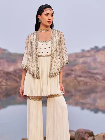 This ivory sharara set features a beautifully embellished peplum top and a delicate cape. The pleated pants add a modern touch, making it a sophisticated choice for any festive occasion.