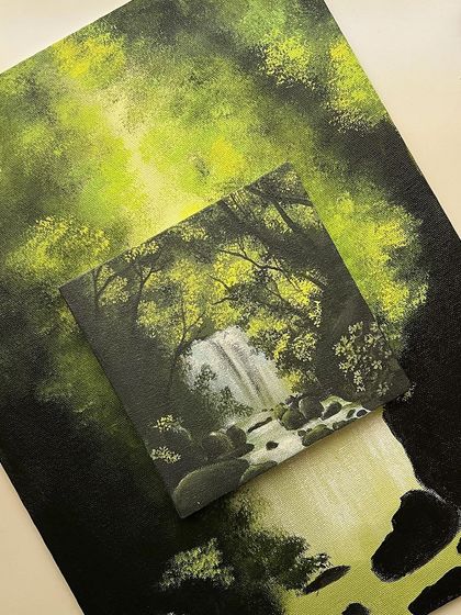 A small, finished waterfall painting placed on top of a larger, unfinished version of the same scene. Sometimes, a small study can provide the motivation to complete a bigger project.