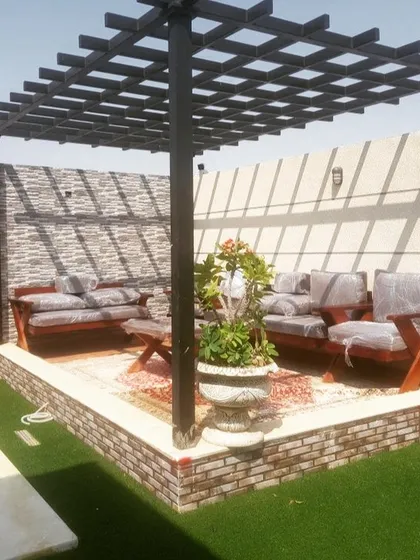 A traditional black pergola with a grid-style roof, providing a classic framework for an outdoor seating area. The base is finished with brick and stone, complementing the artificial grass surround.