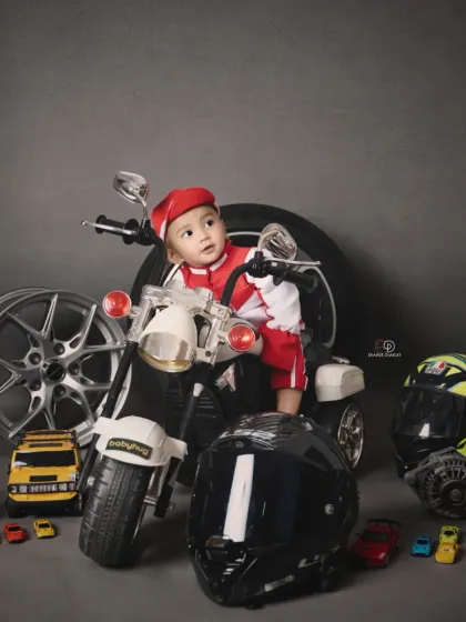 A toddler enjoying his biker-themed photoshoot, sitting on a miniature motorcycle. The set is complete with helmets, wheels, and toy cars for an authentic motor-themed portrait.