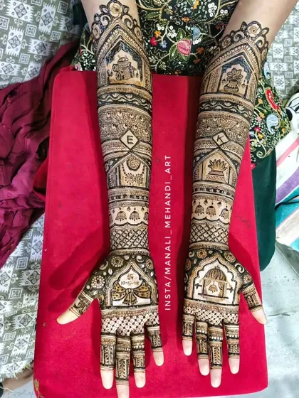 A full-arm bridal design featuring temple gopuram architecture at the top. The arms are filled with various bands of geometric patterns and a personalized initial 'E' for the bride.