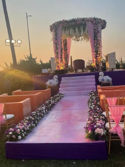 A beautiful sunset view of a wedding mandap set up on a lawn overlooking the sea. The purple platform and orange seating add a pop of color to the romantic evening ambiance.