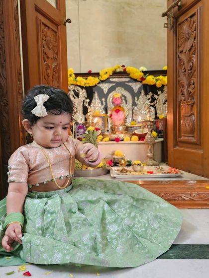 An adorable baby girl in a custom brocade lehenga set in pastel green and peach, sitting by the family pooja mandir.