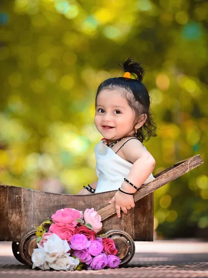 A baby girl in a wooden wheelbarrow filled with flowers, smiling sweetly in a beautiful garden.