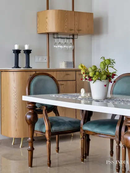 This dining area combines classic and modern elements. Traditional chairs with teal velvet upholstery surround a simple white tabletop, while a light wood bar cabinet with a built-in wine glass rack adds functionality.