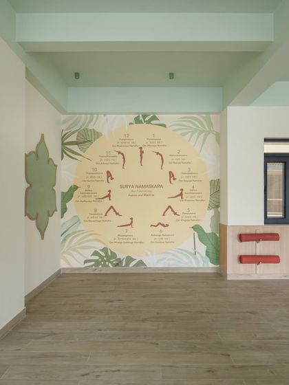 The serene yoga room, featuring a large "Surya Namaskara" mural that guides students through the sequence. The natural wood flooring and leafy graphics create a calm, grounded atmosphere for mindfulness and movement.