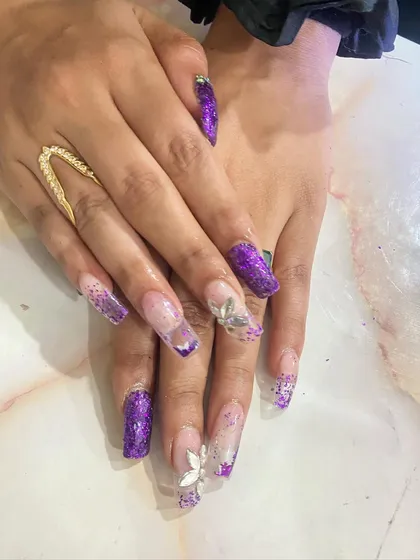 A dramatic purple glitter set on long coffin nails. The design incorporates clear nails with encapsulated purple glitter and silver 3D flower accents.