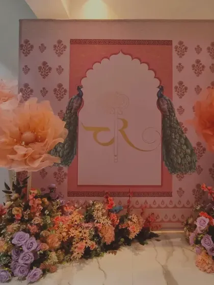 A traditional Indian-themed backdrop perfect for a sangeet or mehendi. It features a Rajasthani arch design with peacock motifs and is decorated with large, colorful paper flowers.