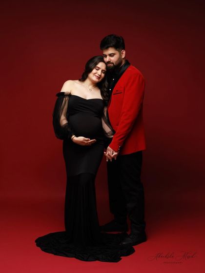 A romantic and elegant maternity portrait. The couple holds hands, sharing a quiet moment of connection against a rich, passionate red backdrop.