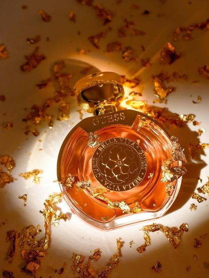 Gold. A luxurious close-up of a Guess perfume bottle, showered in gold flakes to emphasize its opulent fragrance and beautiful packaging. The lighting creates a warm, radiant glow.