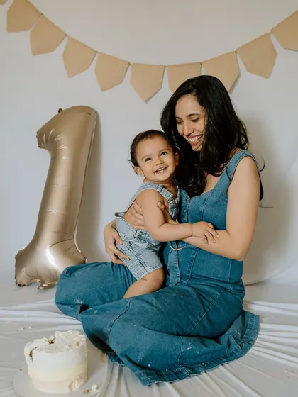 A simple and sweet first birthday moment. The mother holds her smiling baby, both dressed in matching denim, with a "1" balloon and a cake to mark this special occasion.