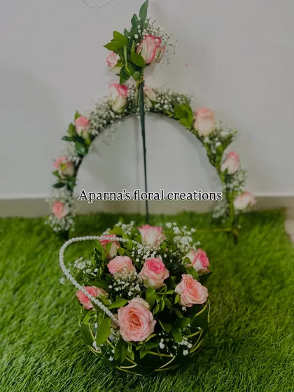A matching set of baby shower props: a floral Dhanushbaan (bow and arrow) and a flower basket. Both are decorated with delicate pink roses and baby's breath.