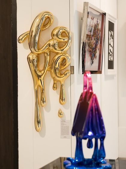 The golden 'Love Drip' sculpture by Sanuj B. installed on a gallery wall. This piece represents love as a fluid, ever-flowing emotion, like a golden liquid that spills over and permeates everything it touches.