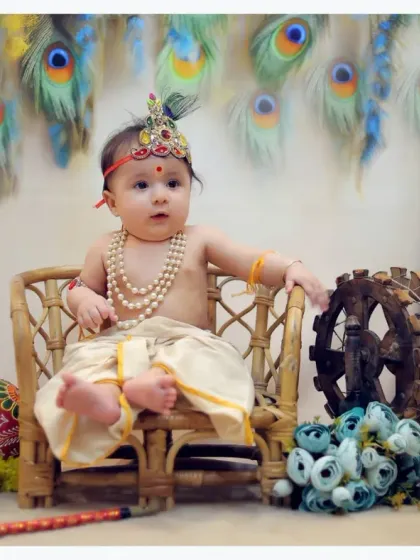 A variation of the Krishna theme, this shot features a baby sitting on a small stool with a spinning wheel prop. The background combines peacock feathers and marigold flowers for a vibrant look.