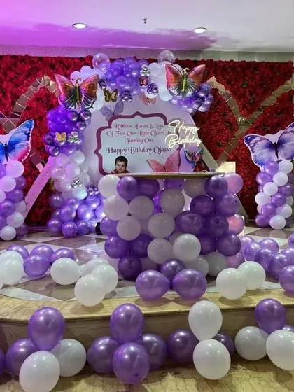 A butterfly-themed birthday stage with a red flower wall backdrop and purple and white balloon columns.