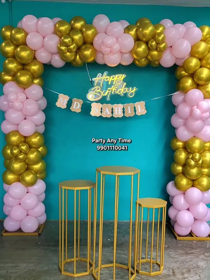 A simple outdoor birthday setup with a pink and gold balloon arch against a teal wall.