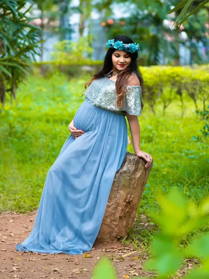 A whimsical outdoor portrait. The mom-to-be sits on a rock in a flowing blue gown, looking like a fairytale character in an enchanted forest.