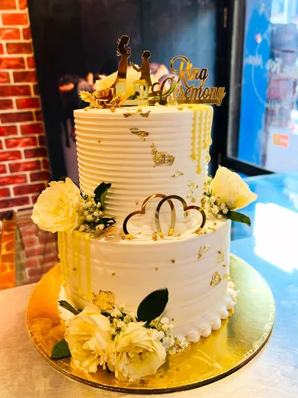 An elegant two-tier Roka ceremony cake in white and gold. It's decorated with fresh white roses, baby's breath, and gold leaf for a touch of luxury.