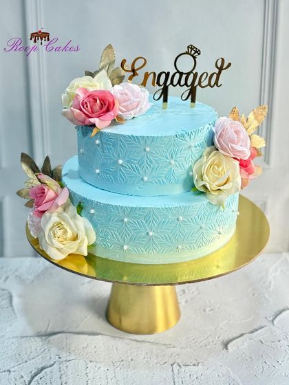A beautiful two-tier engagement cake in a lovely shade of blue. The design features a delicate stencil pattern, pearl details, and is adorned with bouquets of pink, white, and yellow roses.