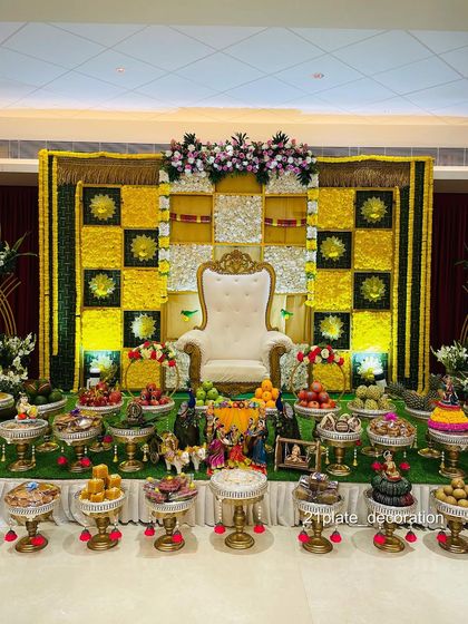 A grand and traditional baby shower stage with a yellow and green checkered floral backdrop. The extensive display of decorated plates and props makes it a complete and auspicious setup.