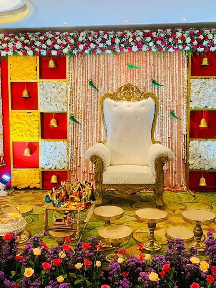 A festive and colorful Seemantham stage with a backdrop of red, yellow, and white panels. The decor includes hanging bells and parrot motifs, creating a vibrant and joyful atmosphere.