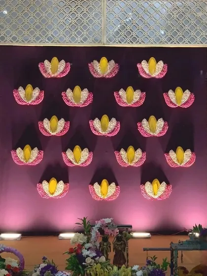 A unique Ganpati backdrop featuring a deep purple wall adorned with lotus flower motifs made from petals. The design is framed by hanging pearl strings, creating a royal and elegant look.