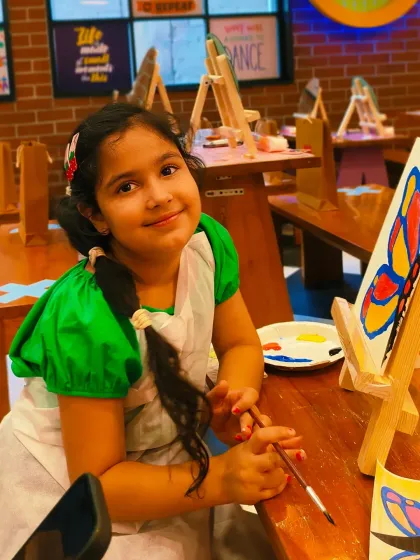 A young artist smiles proudly next to her butterfly painting. Our workshops provide all the materials and a supportive environment to let creativity flourish.