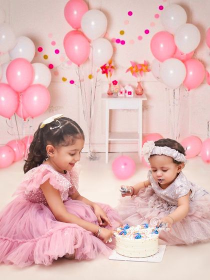 A shared cake smash between sisters is double the fun. The older sister helps the younger one explore her first birthday cake in this sweet, balloon-filled scene.