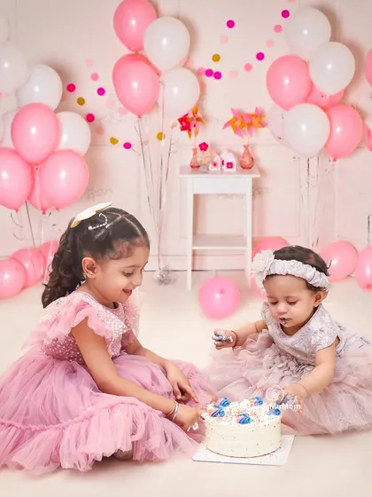 A shared cake smash between sisters is double the fun. The older sister helps the younger one explore her first birthday cake in this sweet, balloon-filled scene.