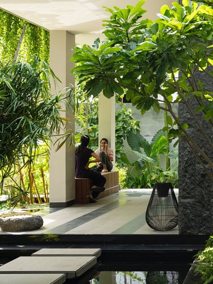 A quiet conversation nook on a covered veranda, looking out onto a water body and garden. The design uses built-in wooden seating and is surrounded by greenery, creating a space that feels both sheltered and open.