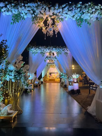 A dreamy and elegant wedding entrance tunnel created with flowing white drapes, white floral arrangements, and sparkling crystal chandeliers. The blue uplighting adds a magical, ethereal glow.