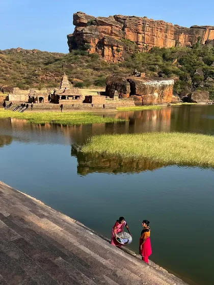 Two women in colorful sarees wash clothes by the river, with ancient temples and rocky hills in the background. This image beautifully blends the daily life of today with the history of the landscape.