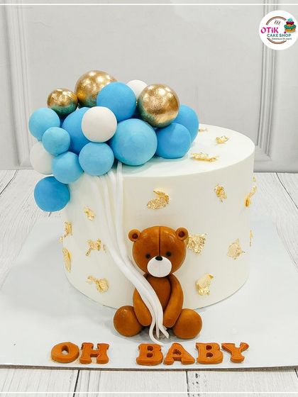 A classic "Oh Baby" cake featuring a cute teddy bear holding a string of blue and gold balloons. The gold leaf accents add a touch of elegance.