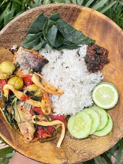 A simple, wholesome farm-style meal. This wooden bowl holds a comforting mix of rice, fish curry with bitter eggplant and foraged greens, and a side of fresh cucumber and lemon.