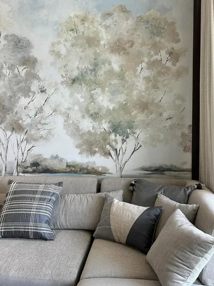 A closer look at the artisanal wallpaper reveals its meticulous craftsmanship and painterly detail. The soft, blended tones of the trees create a calming and inviting atmosphere, perfectly complementing the neutral-toned sofa and cushions.
