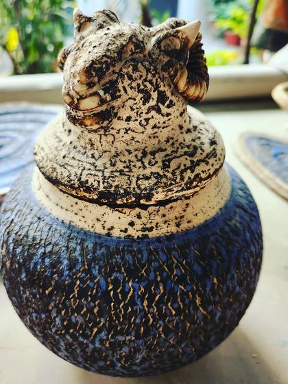 This decorative jar was finished using an Obvara firing technique, which creates the distinctive crackled and colored surface. The lid is sculpted to resemble a ram's head, adding a powerful artistic detail.
