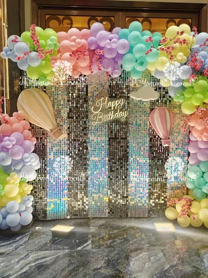 A beautiful hot air balloon themed photo booth. The backdrop is a shimmering sequin wall framed by a pastel rainbow of balloons, with hot air balloon and cloud cutouts adding to the dreamy sky theme.