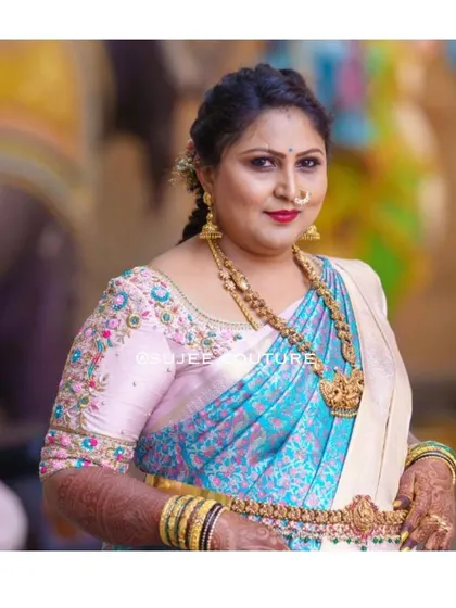 A beautiful client wearing a custom-designed blouse for a family wedding. The light pink blouse features colorful floral threadwork embroidery on the sleeves, perfectly matching her turquoise and cream silk saree.