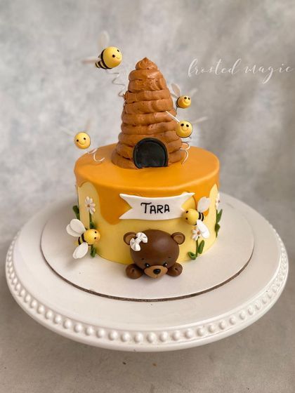What's the buzz? This sweet-as-honey bee-themed cake is decorated with buttercream and features a cute teddy bear, a beehive, and little buzzing bees with delicate wafer paper wings.